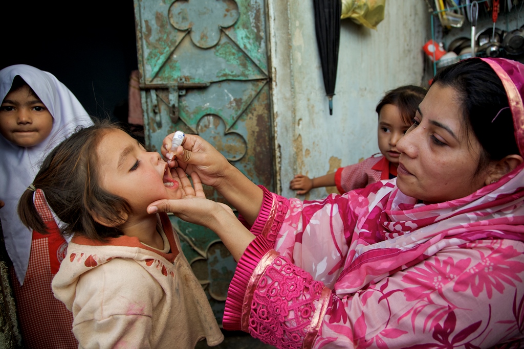 A health worker vaccinates a child against polio outside her home during Polio – National Immunisation Day (NID, while her sisters watching in Sherpow in Landhi town, Karachi Pakistan.